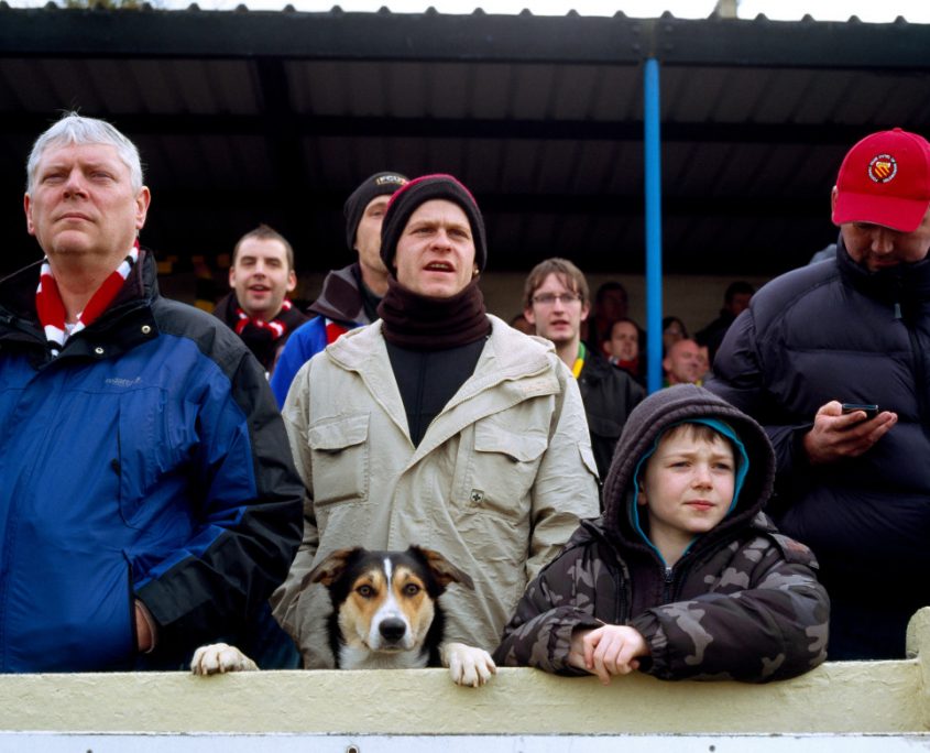  a crowd of people a dog supporting FC United of Manchester peers over the pitch perimeter at Buxton Football Club England